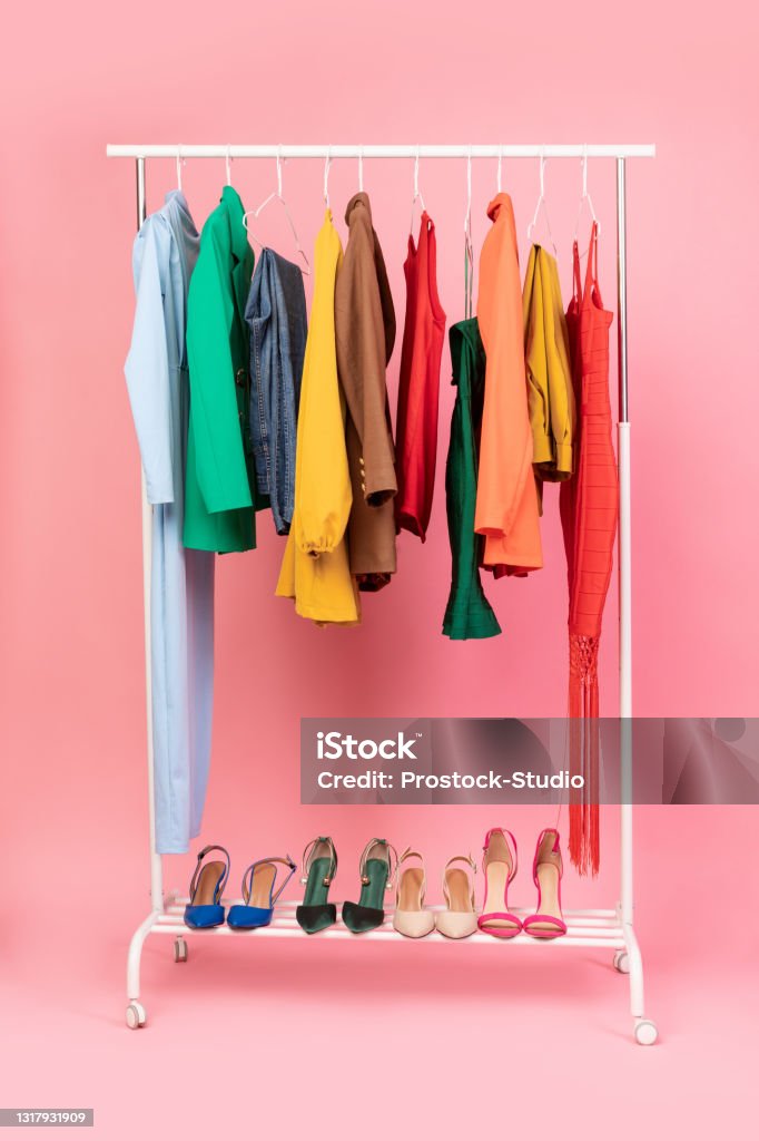 Vertical Shot Of Clothing Rail With Clothes Over Pink Background Shopping Concept. Vertical Shot Of Clothing Rail With Bright Trendy Clothes Hanging On Hangers And Shoes Over Pink Studio Background In Empty Room. Fashion Trends And Style Concept Clothes Rack Stock Photo
