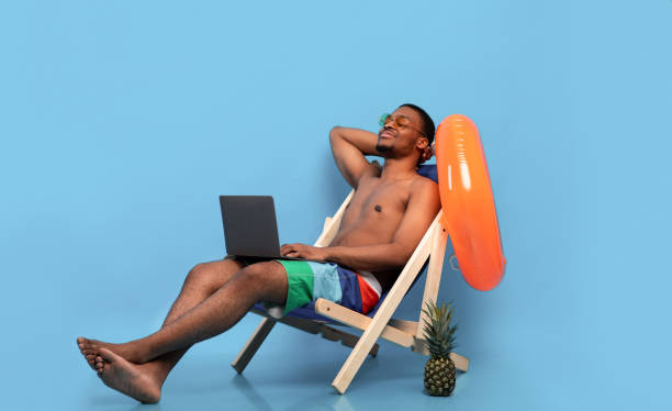 istock Relaxed black guy chilling in lounge chair with laptop computer, working online from tropical paradise, blue background 1317935003