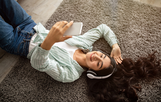 Happy Indian lady in headphones lying on floor and listening to music on mobile device at home, above view. Sweet young woman enjoying her favorite playlist or audio book