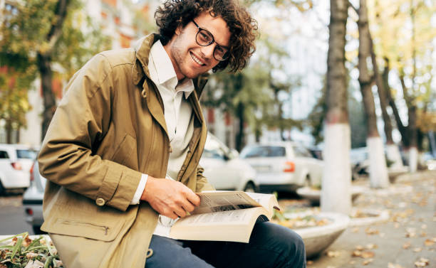 야외에서 책을 읽는 젊은 남자의 수평 이미지. 대학 남성 학생은 가을 거리에서 캠퍼스에서 배운다. 곱슬 머리를 가진 미소 똑똑한 남자는 도시에서 안경과 독서 책을 착용합니다. - men reading outdoors book 뉴스 사진 이미지