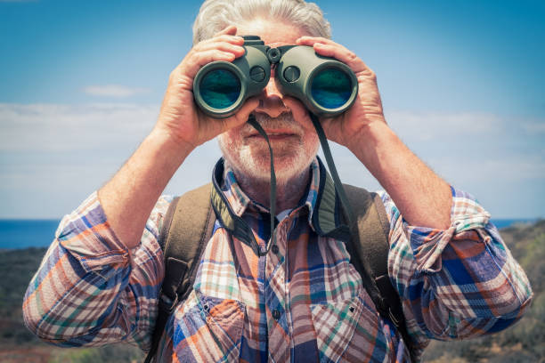 istock Front view of senior white-haired man hiking in outdoors looking with binoculars at camera. Horizon over sea 1317939465