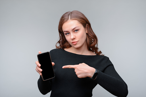 Portrait of a nice young brunette woman holding smartphone with blank screen and pointing at it with her left index finger. Studio shot, gray background