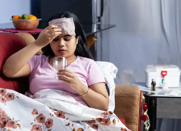 An Indian Asian woman suffering from fever drinking water and applying wet cloth on forehead while lying on sofa