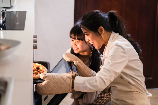 asiatische junge mutter und tochter machen pizza zu hause. frau öffnen sie den ofen und bringen sie das essen aus der maschine. kleines mädchen, das auf das essen schaut und es mit lächelndem gesicht riecht, genießen sie die gemeinsame aktivität. - two parent family indoors home interior domestic kitchen stock-fotos und bilder