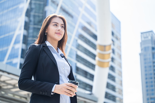 Asian young office businesswoman stand outdoor in city with confident face, enjoy life holding a cup of hot coffee. Working woman feeling fresh and happy with smile after drink refreshment beverage.