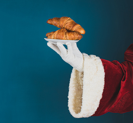 Santa Claus offered croissants. Hand of Santa on a dark turquoise background with copy space.