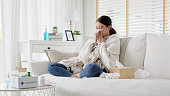 ソファの毛布の下に座り、自宅でティッシュペーパーでくしゃみをする病気の若いアジア人女性。インフルエンザに苦しんで、自宅の組織で鼻を吹いたり、咳やくしゃみをしたりする女性。�