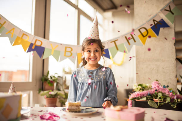 mädchen, das am tisch steht und mit einem zuschüchternden lächeln in die kamera schaut - balloon child people color image stock-fotos und bilder