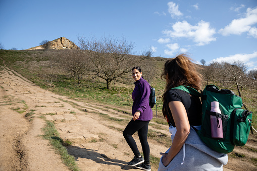Two female friends walking up a footpath trail on Roseberry Topping, Middlesborough, North Yorkshire. One woman is looking back at her friend and talking to her.