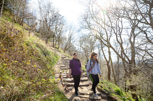 A wide shot of two female friends walking down steps on a footpath trail on Roseberry Topping, Middlesborough, North Yorkshire. They are talking to each other as they walk. The sun is shining above them.