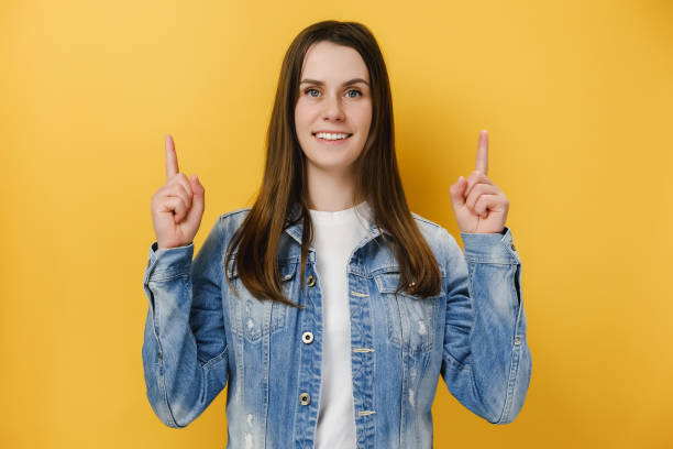 istock Cute young satisfied woman 20s points index fingers up, shows space on yellow studio background wall for your advertisement, wears blue denim jacket and t-shirt, smiles happily, promots place upstairs 1317954300