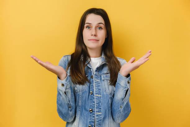 istock Clueless hesitant brunette woman spreads palms sideways stands doubtful cannot make decision, wears blue denim jacket, isolated over yellow studio background. European questioned female has no idea 1317955365