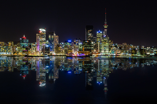 This November 2019 long-exposure image taken at night shows the skyline of Tāmaki Makaurau Auckland.  This image was taken from Cyril Bassett Lookout in Stanley Point. The Sky Tower, an icon of Aotearoa New Zealand, appears prominently. The light trail of a ferry is seen on the right. The original photo was edited to add a reflection of the city skyline in Waitemata Harbour.