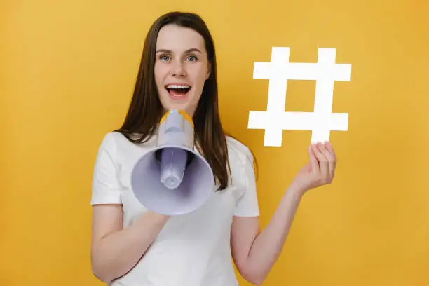 Photo of Surprised happy young girl 20s screaming in megaphone showing white hashtag sign, wears t-shirt, isolated on yellow background. Marketing network, SMM business, customer attraction campaign concept