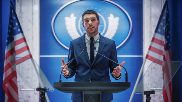 istock Young Organization Representative Greeting Supporters at a Government Election Rally. Press Officer Delivering a Speech at Summit. Minister Speaking to Congress. Backdrop with American Flags. 1317961662