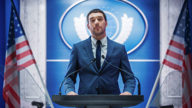 istock Young Organization Representative Greeting Supporters at a Government Election Rally. Press Officer Delivering a Speech at Summit. Minister Speaking to Congress. Backdrop with American Flags. 1317961695