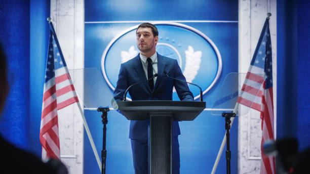 istock Young Organization Representative Answering Questions from Journalists at Press Conference. Press Officer Working at Summit Meeting. Minister Speaking to Congress. Backdrop with American Flags. 1317961708