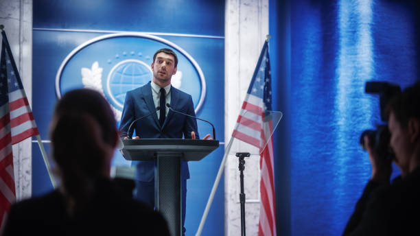 istock Young Organization Representative Answering Questions from Journalists at Press Conference. Press Officer Working at Summit Meeting. Minister Speaking to Congress. Backdrop with American Flags. 1317961714