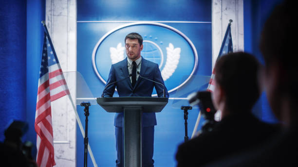 istock Young Organization Representative Answering Questions from Journalists at Press Conference. Press Officer Working at Summit Meeting. Minister Speaking to Congress. Backdrop with American Flags. 1317961716