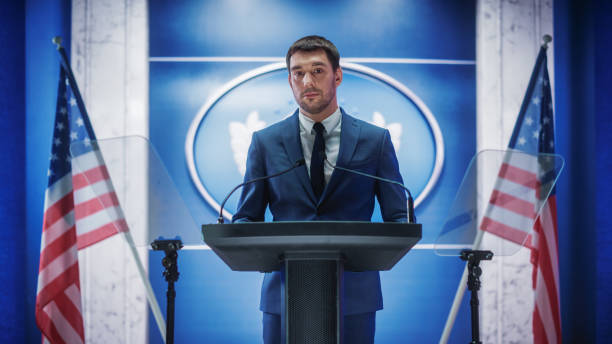 istock Young Organization Representative Answering Questions from Journalists at Press Conference. Press Officer Working at Summit Meeting. Minister Speaking to Congress. Backdrop with American Flags. 1317961724