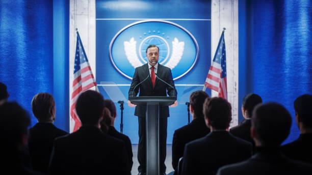 istock Charismatic Organization Representative Speaking at a Press Conference in Government Building. Press Officer Delivering a Speech at a Summit. Minister at Congress. Backdrop with American Flags. 1317961745