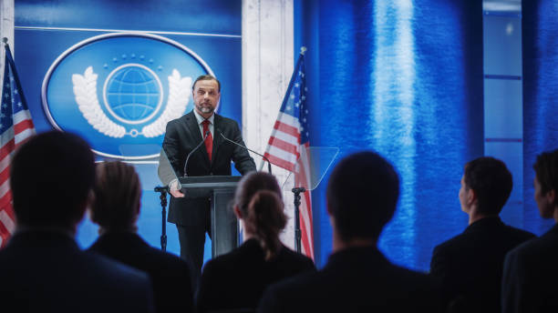 istock Inspiring Organization Representative Speaking at a Press Conference in Government Building. Press Officer Delivering a Speech at a Summit. Minister at Congress. Backdrop with American Flags. 1317961770