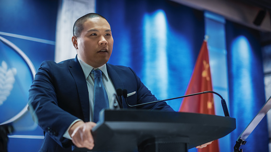 Portrait of Chinese Organization Representative Speaking at Press Conference. Minister Delivering a Speech at Congress. Backdrop with United States of America and People's Republic of China Flags.