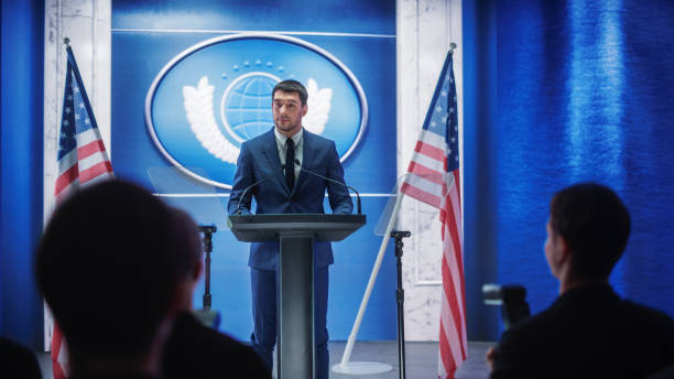 istock Young Organization Representative Answering Questions from Journalists at Press Conference. Press Officer Working at Summit Meeting. Minister Speaking to Congress. Backdrop with American Flags. 1317961794