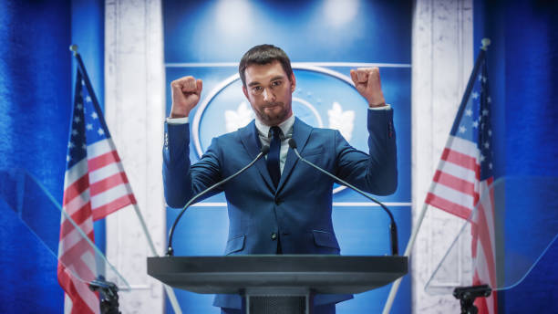 istock Young Organization Representative Delivers a Successful Speech to Supporters at a Government Election Rally, Clenches his Fists. Minister Speaking on Backdrop with American Flags. 1317961798