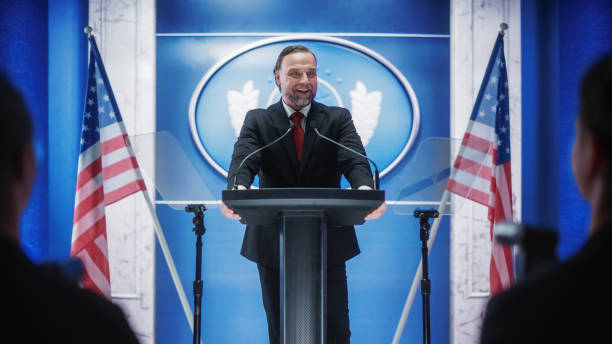istock Charismatic Organization Representative Speaking at a Press Conference in Government Building. Press Officer Delivering a Speech at a Summit. Minister at Congress. Backdrop with American Flags. 1317961801