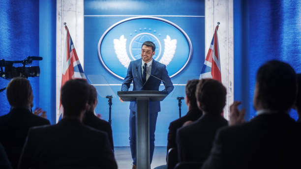istock Young Organization Representative Speaking at Press Conference in Government Building. Press Officer Delivering a Speech at Summit. Minister Speaking to Congress. Backdrop with Great Britain Flags. 1317961819
