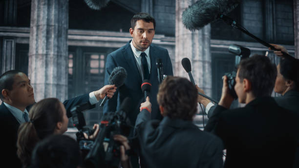 istock Young Organization Representative Answering Press Questions and Giving Interview Outside a Parliament, Court or Other Government Building. Press Officer or Businessman Crowded by News Journalists. 1317961820