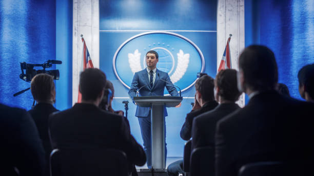 istock Young Organization Representative Speaking at Press Conference in Government Building. Press Officer Delivering a Speech at Summit. Minister Speaking to Congress. Backdrop with Great Britain Flags. 1317961821