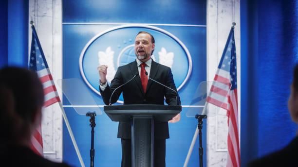 istock Charismatic Organization Representative Speaking at a Press Conference in Government Building. Press Officer Delivering a Speech at a Summit. Minister at Congress. Backdrop with American Flags. 1317961838