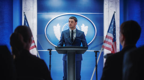 istock Young Organization Representative Answering Questions from Journalists at Press Conference. Press Officer Working at Summit Meeting. Minister Speaking to Congress. Backdrop with American Flags. 1317961842