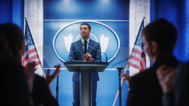 Young Organization Representative Delivers a Successful Speech to Supporters at a Government Election Rally. Crowd is Cheering and Clapping Hands. Minister Speaking on Backdrop with American Flags.