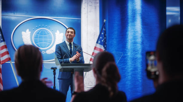 istock Young Organization Representative Delivers a Successful Speech to Supporters at a Government Election Rally. Crowd is Cheering and Clapping Hands. Minister Speaking on Backdrop with American Flags. 1317961873