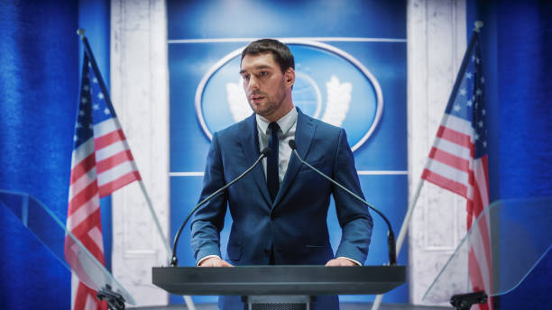 istock Young Organization Representative Answering Questions from Journalists at Press Conference. Press Officer Working at Summit Meeting. Minister Speaking to Congress. Backdrop with American Flags. 1317961885