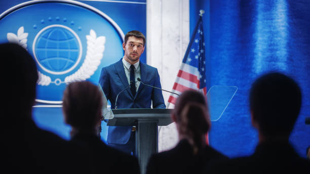 istock Young Organization Representative Answering Questions from Journalists at Press Conference. Press Officer Working at Summit Meeting. Minister Speaking to Congress. Backdrop with American Flags. 1317961910