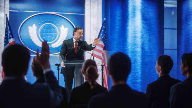 istock Charismatic Organization Representative Speaking at a Press Conference in Government Building. Press Officer Delivering a Speech at a Summit. Minister at Congress. Backdrop with American Flags. 1317961915