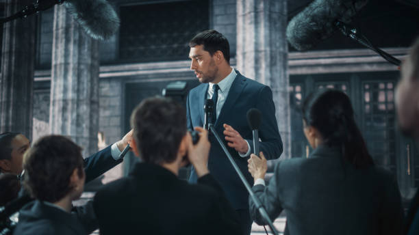 istock Young Organization Representative Answering Press Questions and Giving Interview Outside a Parliament, Court or Other Government Building. Press Officer or Businessman Crowded by News Journalists. 1317961931
