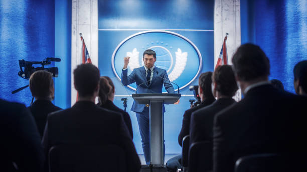 istock Young Organization Representative Speaking at Press Conference in Government Building. Press Officer Delivering a Speech at Summit. Minister Speaking to Congress. Backdrop with Great Britain Flags. 1317961937
