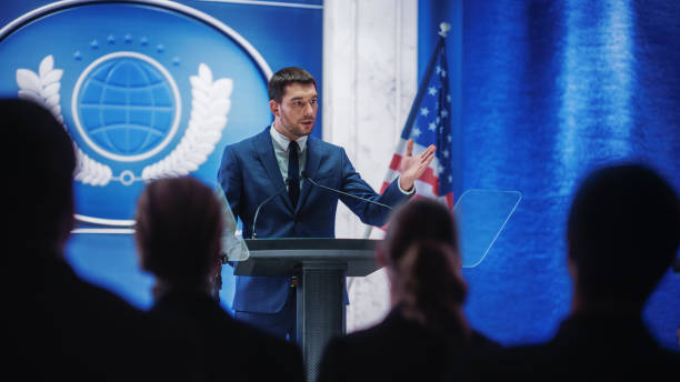 istock Young Organization Representative Answering Questions from Journalists at Press Conference. Press Officer Working at Summit Meeting. Minister Speaking to Congress. Backdrop with American Flags. 1317961958