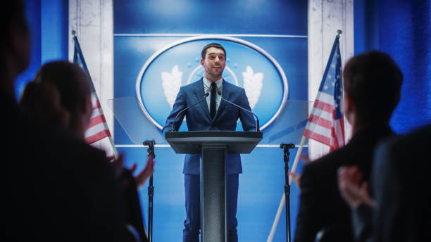 istock Young Organization Representative Delivers a Successful Speech to Supporters at a Government Election Rally. Crowd is Cheering and Clapping Hands. Minister Speaking at Summit. 1317961969
