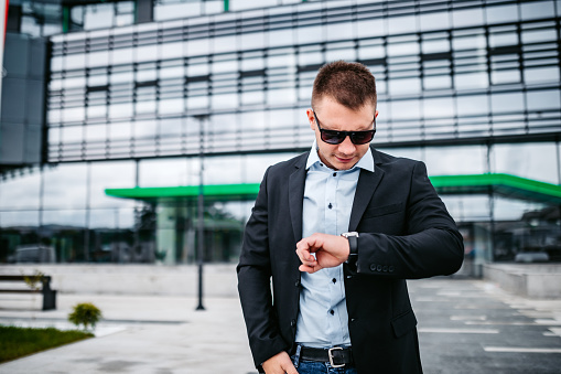 Portrait of young handsome businessman standing in front of business building and checking time on wristwatch.