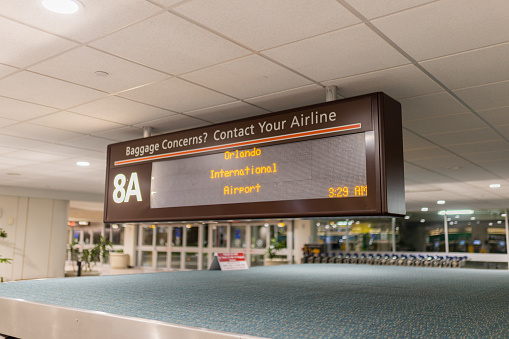 Airport signboard displaying Orlando International Airport in yellow letters. Black digital sign hanging from white ceiling with windows and glass doors as background. Transportation and airplanes