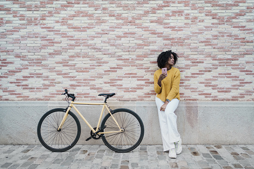 Attractive black woman standing over brick wall next to a modern urban yellow bike, with smart phone on her hand
