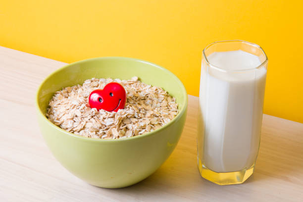 aveia em uma tigela verde com um coração vermelho feliz e um copo de leite em uma mesa de madeira, cuidados de saúde, alimentação e alimentos adequados, tolerância ao glúten e lactose, fundo amarelo - oatmeal breakfast healthy eating cholesterol - fotografias e filmes do acervo