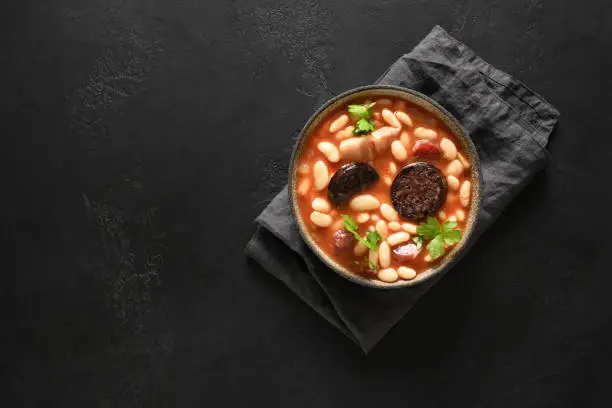 Photo of Spanish asturiana fabada with chorizo and morcilla on black background.