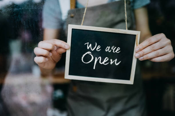 close up of young asian female small business owner in apron starting her business day. hanging an open sign on the door at the shop. business owner and everyday business practice concept - boutique owner store retail occupation imagens e fotografias de stock